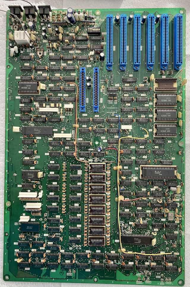The motherboard has been removed from the system. The back of the computer is at the top of the picture, with six blue expansion card slots. There are two smaller expansion slots marked "MEM" near the middle, and a sea of 74 DIP logic along with a 6845, 6809, and 6821.