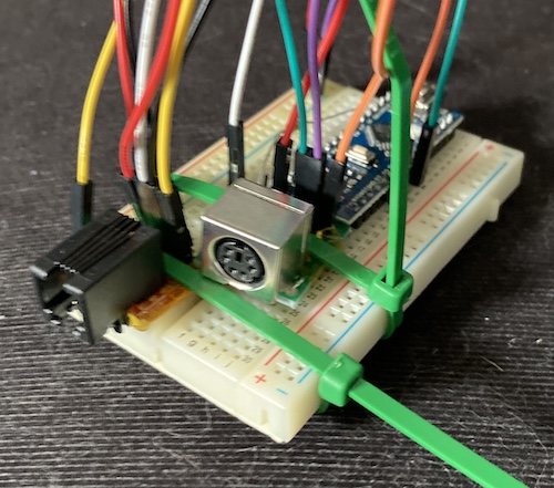 A solderless breadboard with a PS/2 breakout and a 4P4C breakout ziptied to it. An Arduino Mini knockoff board is installed into the breadboard and a maze of wires goes everywhere.