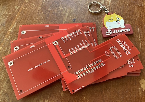 A bunch of red Z-shaped power supply boards, along with a cute dog keychain that reads "JLCPCB." They are sitting on a battered kitchen table that sorely needs resurfacing; check in at the Petrified Of Wood project blog to see how that went.
