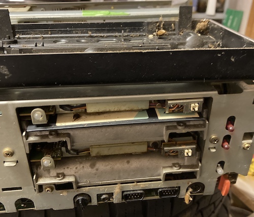 The front fascia of the computer is removed, and placed on top. You can see lots of gunk inside it, as well as left behind on the metal case. A dead bug is in the foreground, dangling from a long-dead spider's web.