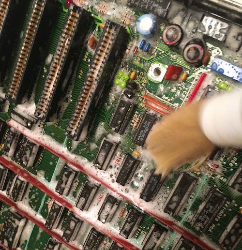 Soap suds are all over the logic board, along with a boar hair brush agitating them in the foreground.