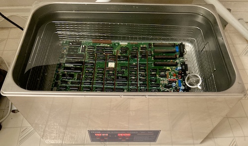 The Apple III logic board is immersed into the basket of a shiny stainless steel ultrasonic cleaner.