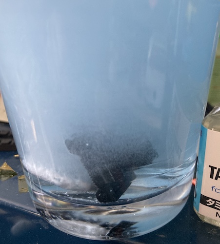 Blue and white fizzy liquid surrounds the black plastic sliders that are stuck at the bottom of this glass cup full of hot Polident.