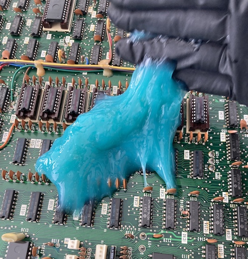 The blue ColorCoral slime is being rolled around the motherboard by my gloved hand. It is sticking to a lot of bypass capacitors and ICs.