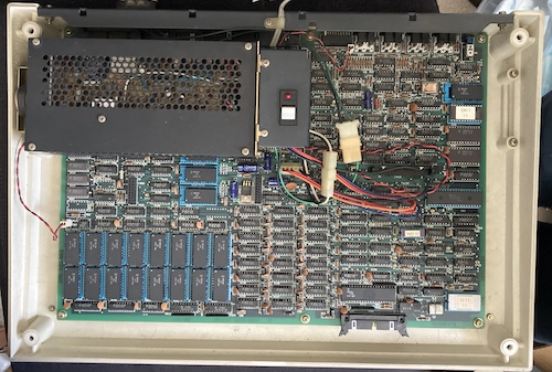 The motherboard is exposed. The black power supply lies over top of most of the ICs in the top left, and socketed ROMs lie in the lower left. The lower right seems to mostly be decoding logic and the keyboard header, and the upper right holds the sub-CPU as well as many other socketed chips, likely gate arrays.