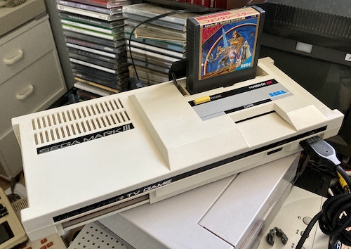 Phantasy Star is inserted into a running Mark III. The power LED is on. There is a pile of junk in the background.
