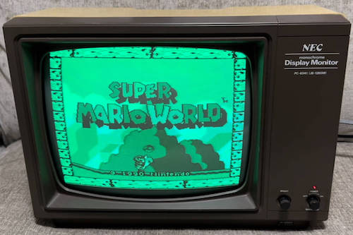 An NEC PC-6041/JB-1260M showing Super Mario World's title screen. It has a black/dark brown casing and says "NEC monochrome Display Monitor" but is otherwise identical
