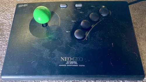 My worn-out Neo Geo joystick: the surface is scratched and covered in dust and cat hair, the logo silkscreen is almost totally worn away, and the buttons are all pointing out at jaunty angles. Someone (me) has replaced the original cracked joystick ball with a nasty green one.