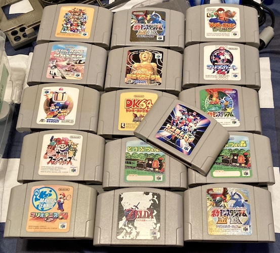 A bunch of Nintendo 64 cartridges lying on the upper shell of an FM Towns IIHR, lying on my guest bedroom's comforter. There are multiple copies of Animal Forest, and multiple copies of some Pokemon game. Custom Robo sits on top, proudly, because there wasn't enough room on the computer to put any more cartridges on.