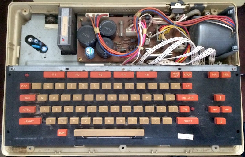 The PC-6001 top case is removed. You can see the power supply board and a very large aluminum heat shield covering everything else of note, other than the keyboard, which is dusty and grimy.