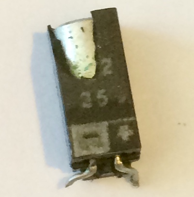 The 14J2 capacitor with its plastic shell broken open. You can see a shiny cylinder inside, dotted with green battery corrosion. The legs are very dull.