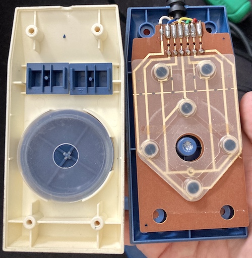 The joystick has been opened. On the left, a clear plastic disc rotates on a blue plastic pad. Two small blue buttons slide in and out of the fascia. On the right, there is a membrane pad with six conductive pads onto traces of a gold-plated PCB.