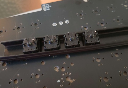 A rigid black plastic plate holds four switches in square alignment, with a spacing lip, for when they're being soldered into the board. This image courtesy of Rayndalf. Thank you!