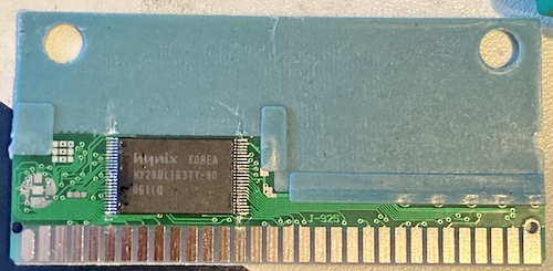 The cartridge PCB is exposed. It has a small circuit board with grey tinned contacts, a TSOP surface-mount flash ROM, and a big, dull grey-blue chunk of something occupying most of the board.
