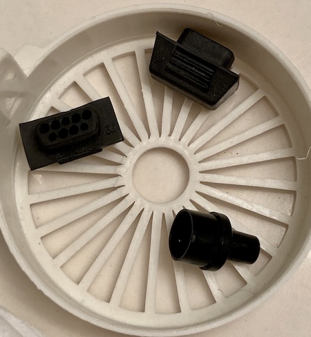 The CMT connector and joystick covers, after cleaning. They are sitting in a white plastic basket meant for holding the things being cleaned. The CMT connector is now obviously clean, shiny plastic.