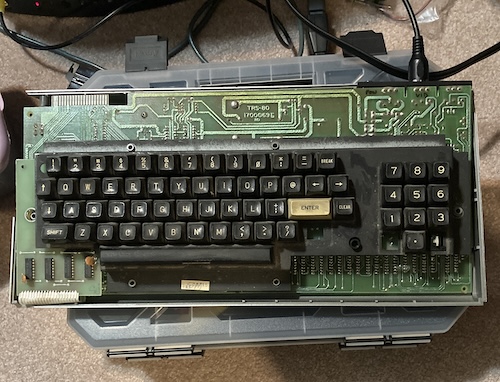 The TRS-80 is sitting on top of some storage containers, plugged into a TV, on carpet.