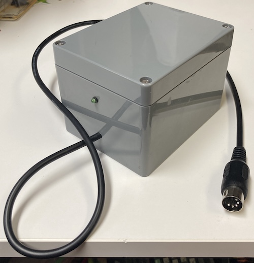 The completed power supply, sadly not powered on at the moment. It's a featureless grey plastic block with a 5-pin DIN cable sticking out of it and a green LED stuck in it.