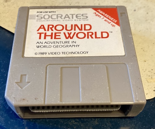 A plastic cartridge, shaped like a floppy disk, reads "For use with Socrates Educational Video System. AWARENESS, Ages 7 and Up, Around The World: An Adventure in World Geography, copyright 1989 Video Technology"