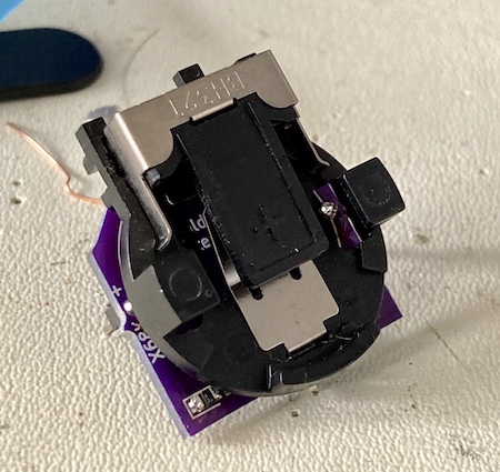 The battery holder PCB assembled. It has a coin-cell battery holder, some legs, and an 0805 surface-mount diode in the corner. A silkscreen marking in the side says "+ TO X68K"
