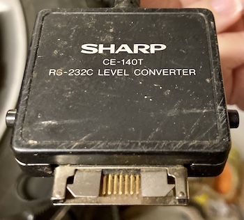 The weird black cable connected to the serial port; it says SHARP CE-140T RS-232C Level Converter. There are gold contacts at the bottom with a strange crablike latch.