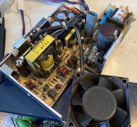The power supply is opened; a grungy 12V fan is in the foreground and you can see a urine-yellow PCB with some caps.